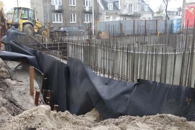 Jedna z faz montażu pionowej izolacji obwodowej fundament&oacute;w: zakładanie i zasypywanie folii hydroizolacyjnej na zewnętrzną powierzchnię ściany fundamentowej.
J. Sawicki